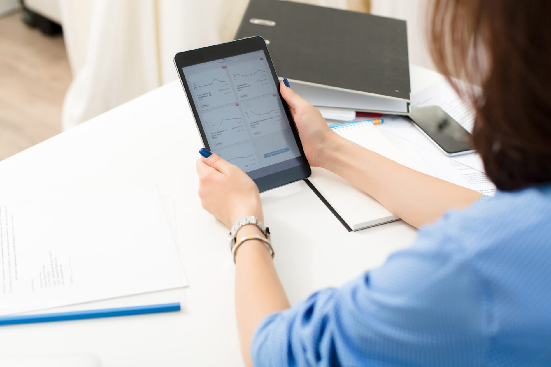 Girl is sitting at the white table in office. She holds tablet and works with financial diagrams.